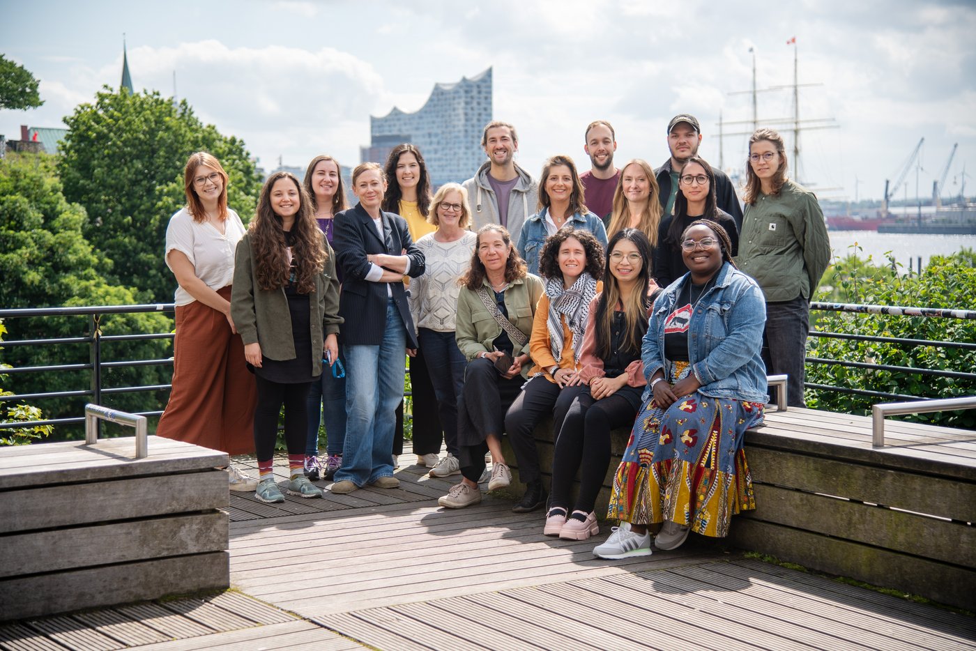 The picture shows a team of 17 people sitting on a bench or standing behind it on a sunny day. In the background you can see the building of the Hamburg Elbphilharmonie and the masts of a sailing ship.