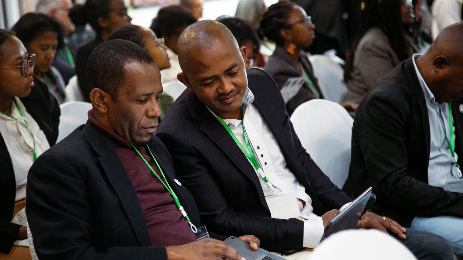 Two CCSM 2024 conference attendees seated together, focused on a laptop screen, wearing event badges with green lanyards in a formal setting.