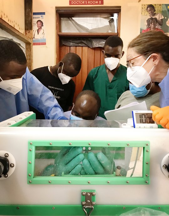Prozedur des "Handschuhwechsels" Drei lokale Labormitarbeitende, ein Ausbilder, der eine persönliche Schutzausrüstung (blauer Kittel und orangefarbene Handschuhe) trägt, und zwei Auszubildende, die ebenfalls eine ähnliche persönliche Schutzausrüstung tragen, stehen um eine Glovebox herum. Ein Auszubildender hat seine Hände in der Glovebox. Er wechselt sicher die Handschuhe, die mit der Glovebox verbunden sind, indem er die alten Handschuhe durch neue ersetzt. Dabei wird er von einem zweiten Auszubildenden unterstützt. Der Ausbilder beaufsichtigt und leitet den Vorgang des Handschuhwechsels an. Die drei anderen Teilnehmer:innen beobachten den Vorgang.