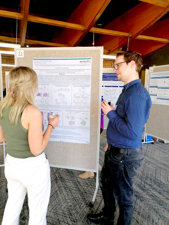 Marco ER-Lukowiak present their latest research results in the poster session of the Gordons Conference. Herr ER-Lukowiak steht neben seinem Poster und diskutiert freundlich mit einer interessierten Frau, die mit dem Rücken zur Kamera steht und sich die Ergebnisse ansieht.