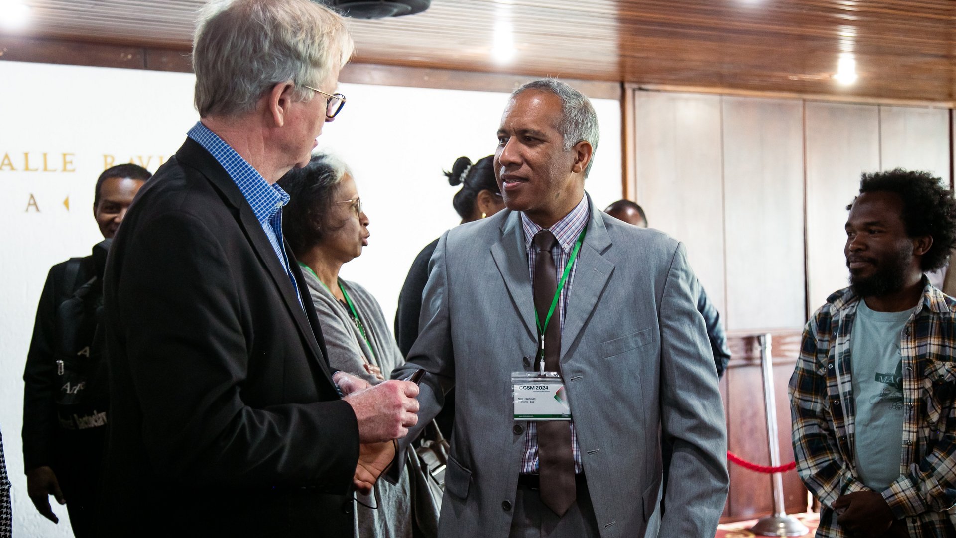 Two men in formal attire engaged in conversation at the CCSM 2024 conference, surrounded by other attendees, with event badges visible.