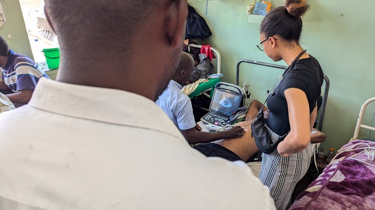 Hands on point-of-care ultrasound (POCUS) training. The image shows a hospital room. There is a person in a white shirt standing in the foreground. In the background, there is a woman wearing a black and white plaid skirt and a black t-shirt. The woman is looking down at a man, who is lying on a bed and receiving a medical examination that involves an ultrasound machine.