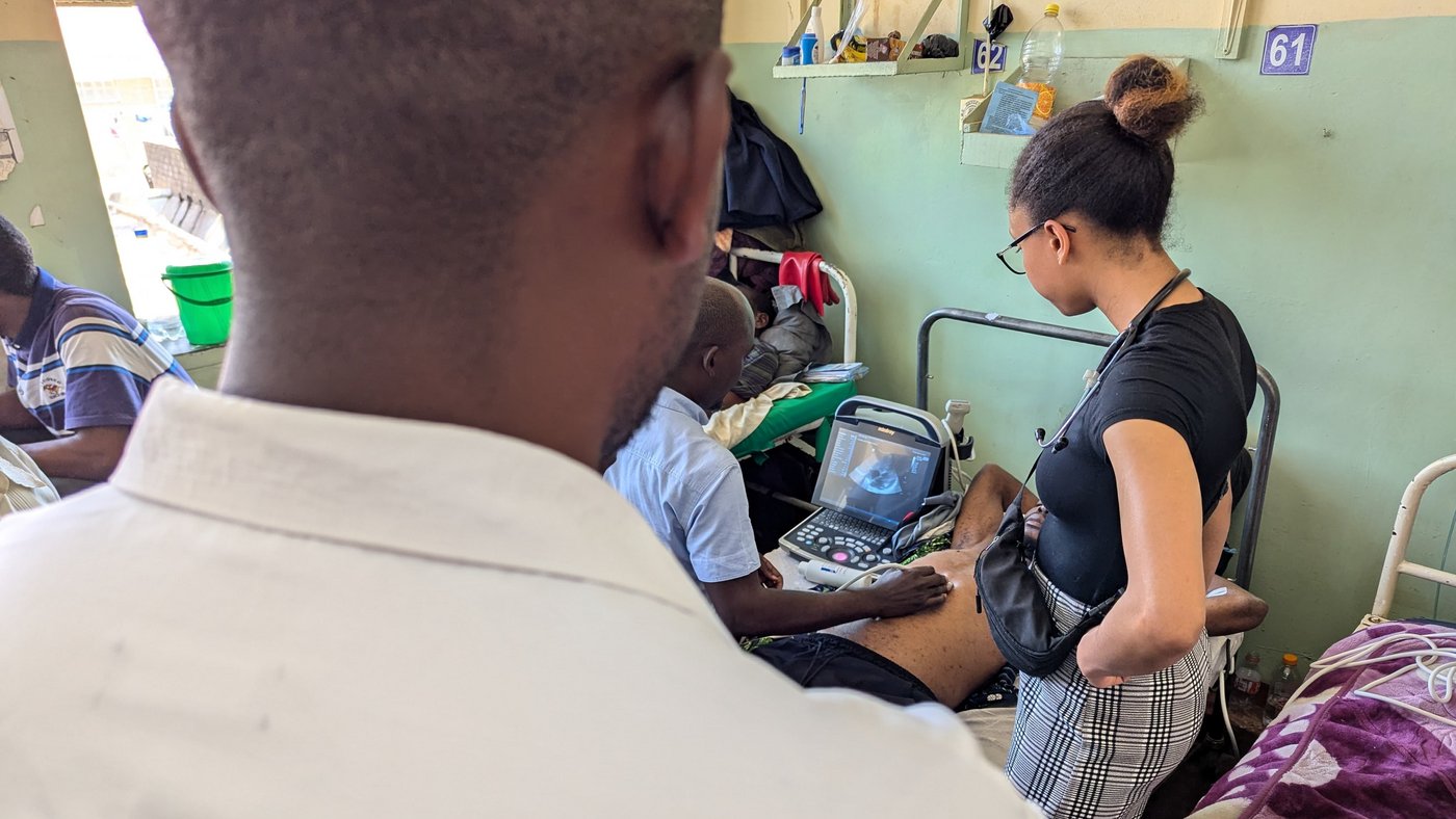 The image shows a hospital room. There is a person in a white shirt standing in the foreground. In the background, there is a woman wearing a black and white plaid skirt and a black t-shirt. The woman is looking down at a man, who is lying on a bed and receiving a medical examination that involves an ultrasound machine.