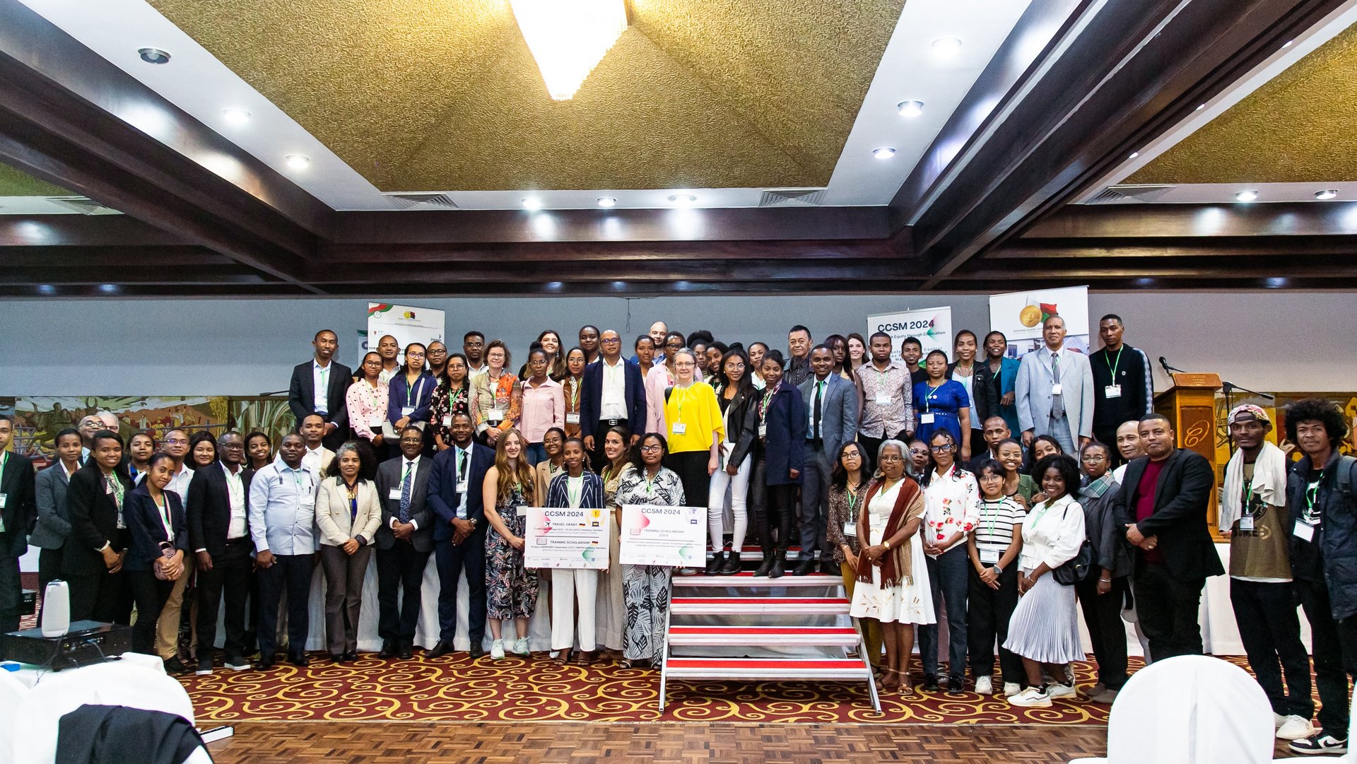 A large group of CCSM 2024 participants posing together on a stage with conference banners and awards displayed in the foreground, under a well-lit ceiling.