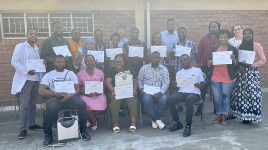 The picture shows the participants and some tutors and organisers of the POCUS train-the-trainers and beginner course at KUHeS in Blantyre, Malawi. The picture shows a group of twenty people standing and sitting in front of a brick building. Some people are holding certificates and ultrasound machines.