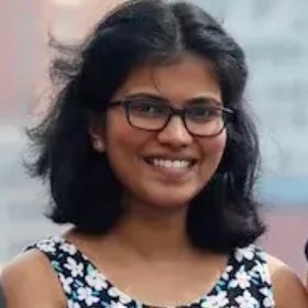 A woman with dark, shoulder-long hair and wearing dark framed glasses smiling kindly into the camera.