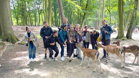 AG Molecular Infection Immunology in the Wildlife Park July 2023. Auf dem Foto steht die Arbeitgruppe in einem Wald des Wildparks Schwarze Berge. Einige Mitglieder füttern vier Rehe, die im Vordergrund zu sehen sind.