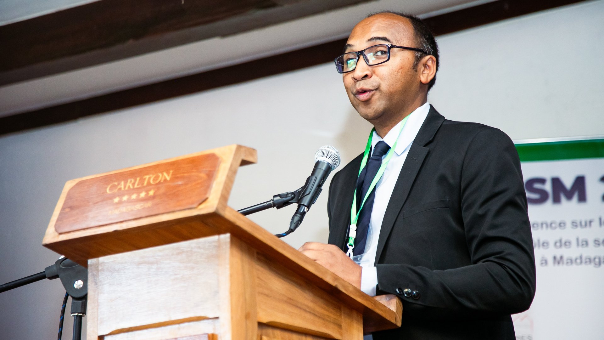 A man wearing glasses and a suit speaking at a wooden podium, addressing an audience at the CCSM 2024 conference.