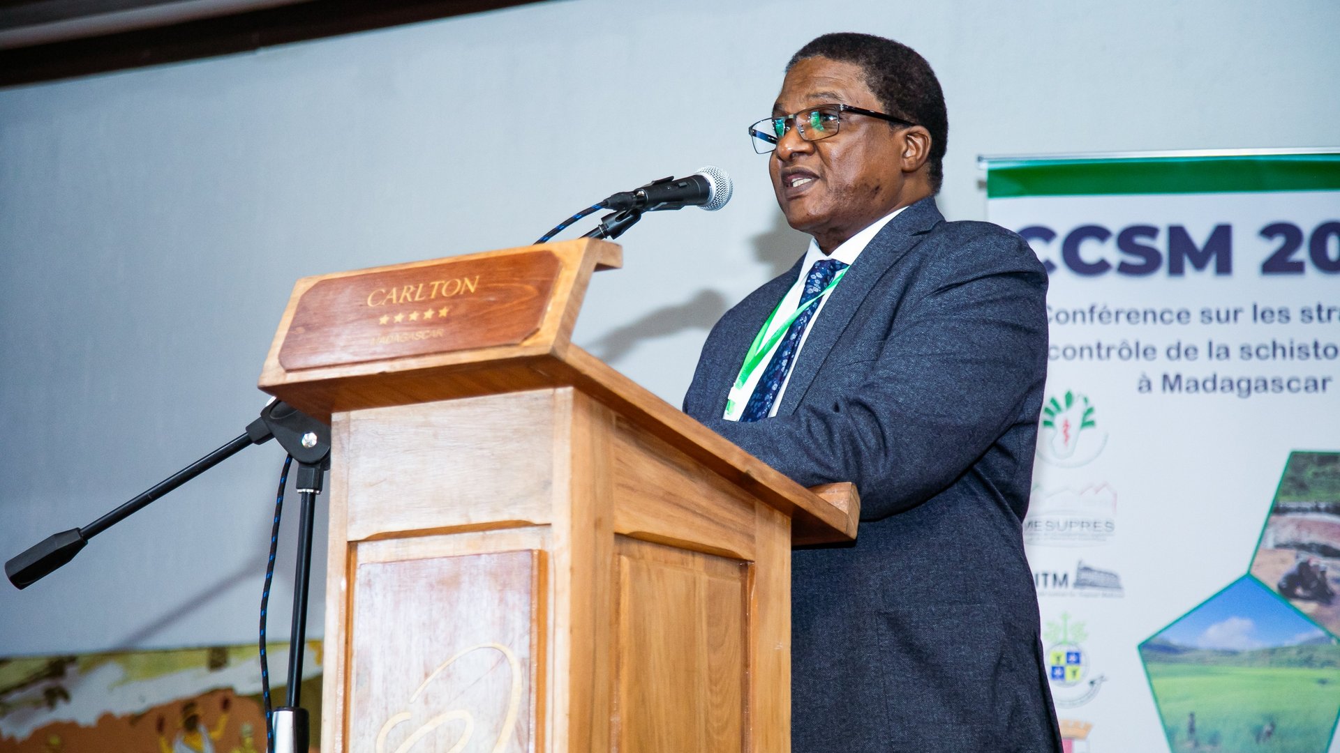 A man in a suit and tie speaking at the CCSM 2024 conference at a wooden podium, with a CCSM 2024 conference banner in the background.