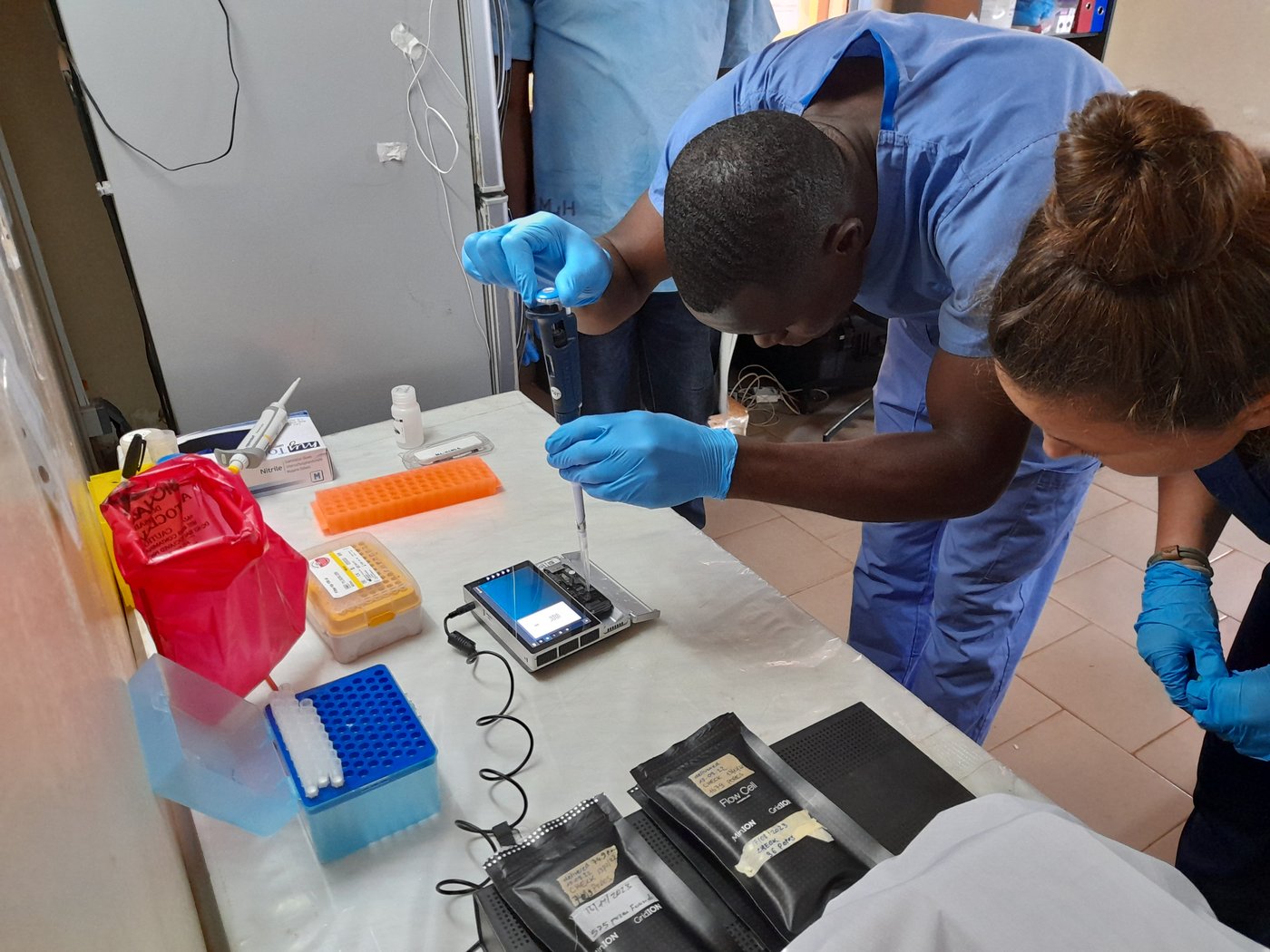 A lab staff is pipetting a sample into a sequencing device, watched by a colleague. He is wearing a blue lab suit and bright blue gloves, red, orange, yellow and blue lab equipment is on the table.