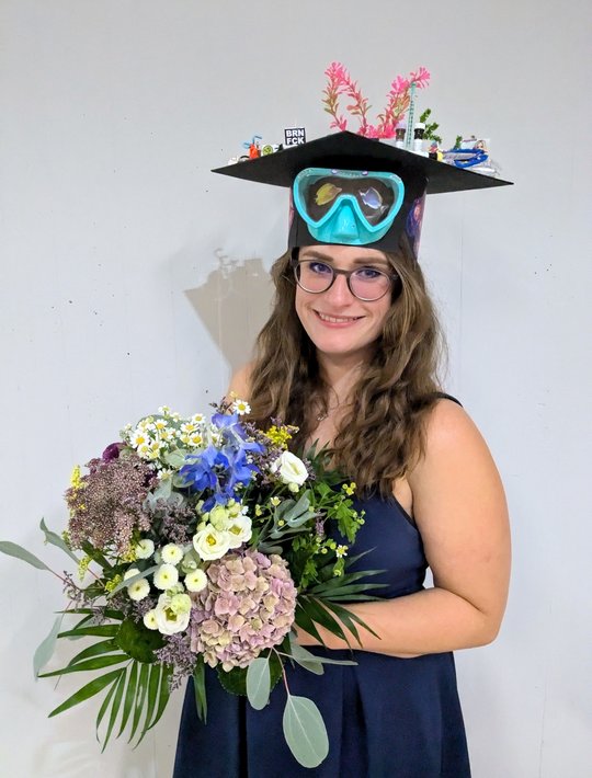 Dr. Charlotte Hansen September 2024 the picture shows charlotte hansen with her doctoral hat and a bouquet of flowers after completing her dissertation. A pair of diving goggles is attached to the hat, which was made by her colleagues, and on top you can see aquarium plants, a syringe and antibody tubes