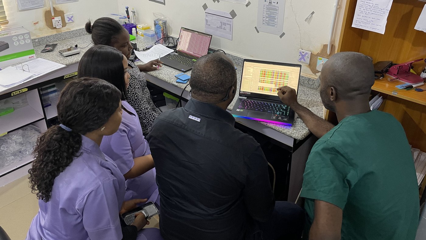 The photo shows five Nigerian colleagues at a lab bench with two laptops on it. They are looking at one of the screens on which a bioinformatic analysis is being performed.