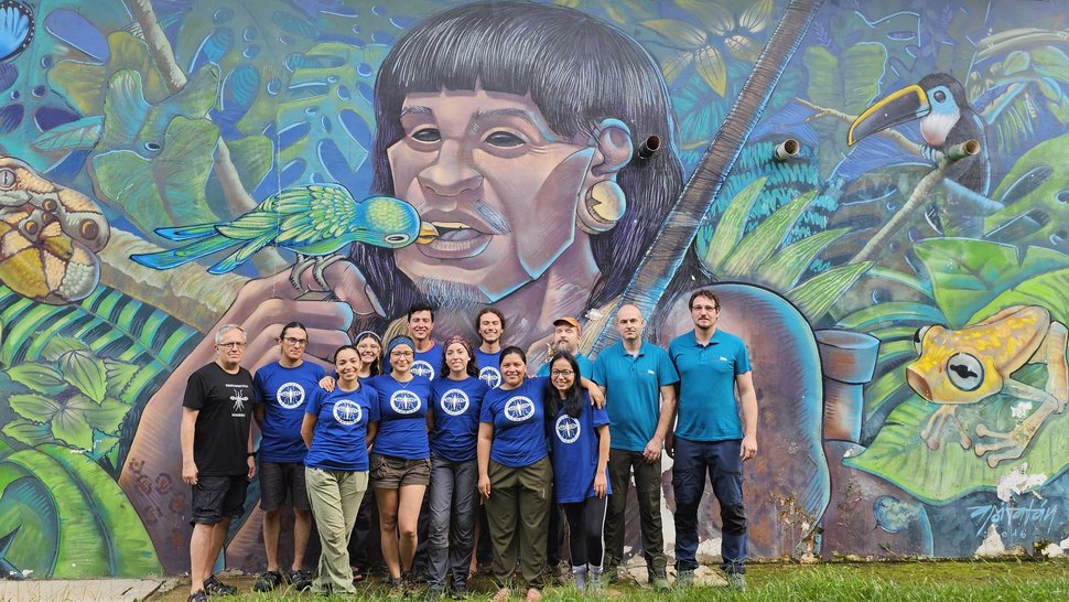 Die AG Virus Metagenomik und Evolution Das Foto zeigt eine Gruppe Menschen vor einem überlebensgroßen bunten Wandgemälde: Ein indigenes Gesicht aus Peru, umgeben von Urwald, in dessen Mund gerade ein Papagei fliegt. Passend zum One-Health-Gedanken.
