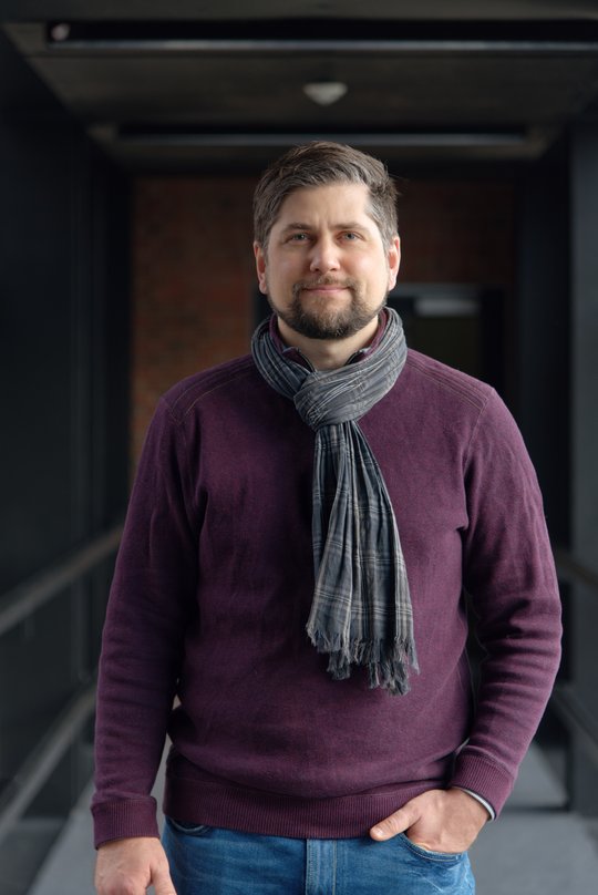 Dr. Johannes Mischlinger Dr. Johannes Mischlinger: ein Gruppenleiter, der kurzes, nach hinten gegeltes braun-graues Haar, einen leichten Bart, einen Schal und ein lila farbendes Oberteil trägt.