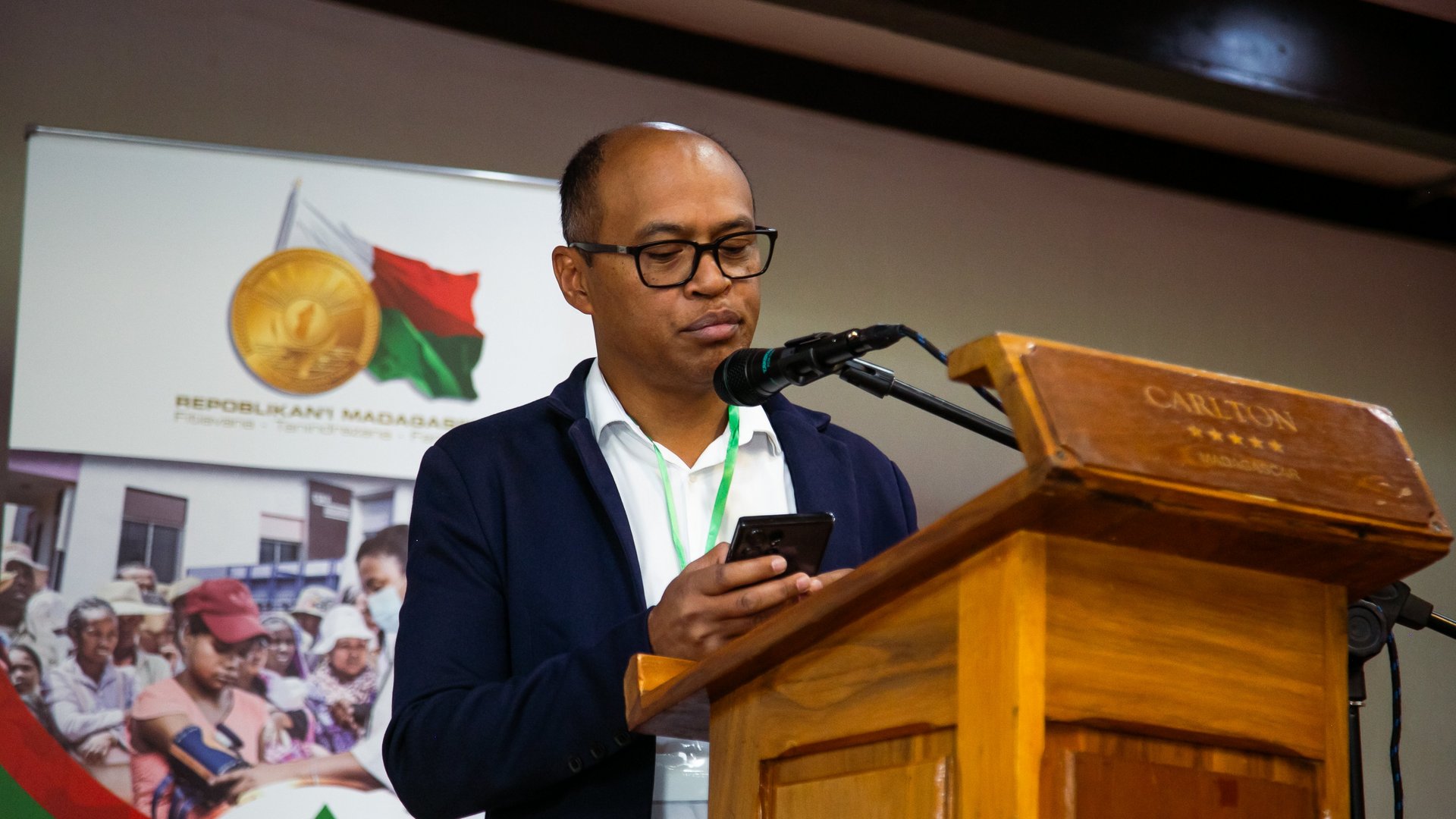 A man speaking at the CCSM 2024 conference at a wooden podium, reading from his phone, with a banner featuring the Madagascar flag and emblem in the background.