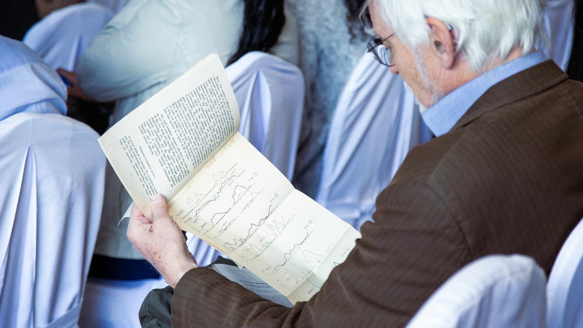 An attendee seated at the CCSM 2024 conference, reading a booklet with graphs and text, wearing a brown jacket in a formal setting.