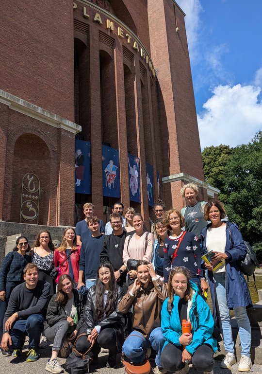 The working groups led by H. Lotter and I. Bruchhaus on their trip to the Hamburg Planetarium In the foreground, a group of 19 people are kneeling and smiling at the camera. In the background, you can see the front of a red brick building with large golden letters spelling out ‘Planetarium.’ Below that are colourful constellations displayed on posters.
