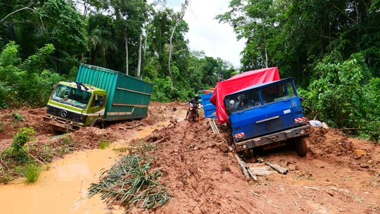 Koloniale Infrastruktur in der DRC Auf dem Foto sind zwei liegen gebliebene Lkws auf matschigen Straßen im Regenwald zu sehen. Eine Person auf einem Motorrad fährt nahe an einem der Lkws vorbei.