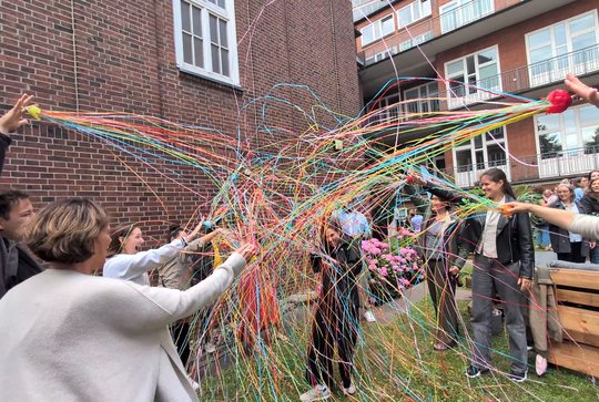 Award winner Barbara Honecker and her colleagues in the BNITM garden Barbara Honecker, who has just been awarded an honour, can be seen here in the garden with her colleagues, who shower her with streamers. She laughs, holds a drink in her hand, and bows her head.