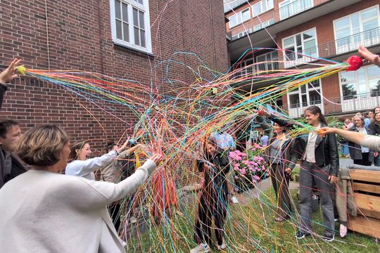 Award winner Barbara Honecker and her colleagues in the BNITM garden Barbara Honecker, who has just been awarded an honour, can be seen here in the garden with her colleagues, who shower her with streamers. She laughs, holds a drink in her hand, and bows her head.