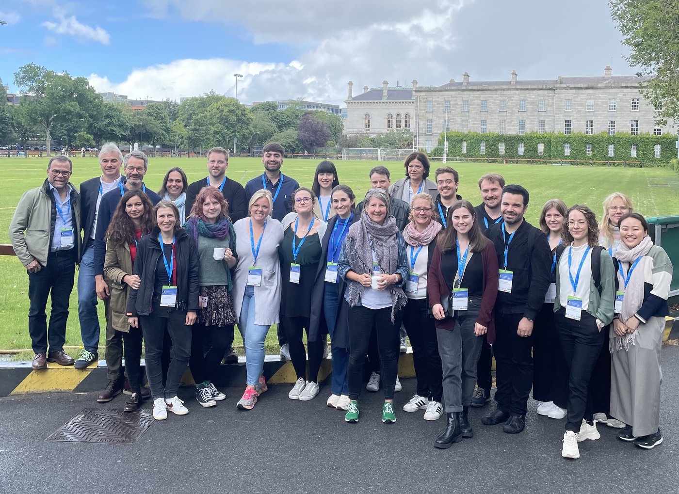 Members of the Sex Differences in Immunity research group stand outside a grassy area and Trinity College in Dublin. They all look cheerful and wear a conference badge around their necks