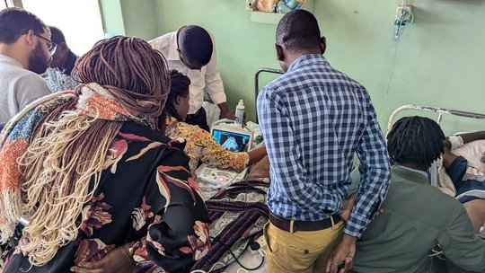 Hands on point-of-care ultrasound (POCUS) training. The picture shows a hands on point-of-care ultrasound training. Several students are gathered around a patient's bed who is being scanned.