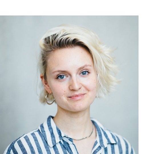 Anna Meisinger Portrait of a white young person with a blonde bob wearing a striped shirt - smiling into the camera and standing against a concrete wall.