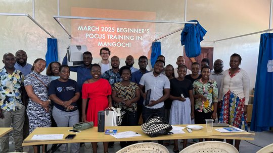 Group picture of the participants and trainers of the POCUS training. The picture shows a group of people standing in front of a wall which says "March 2025 Beginner's POCUS Training in Mangochi"