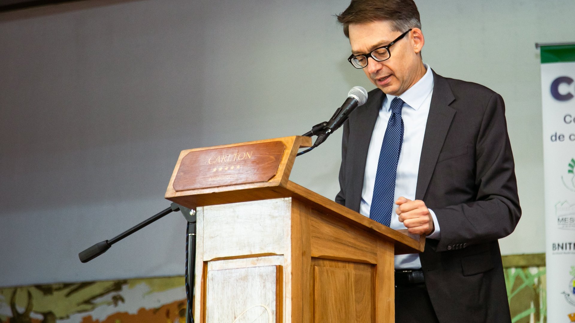 A man in a suit and tie speaking into a microphone at a wooden podium during the CCSM 2024 conference.