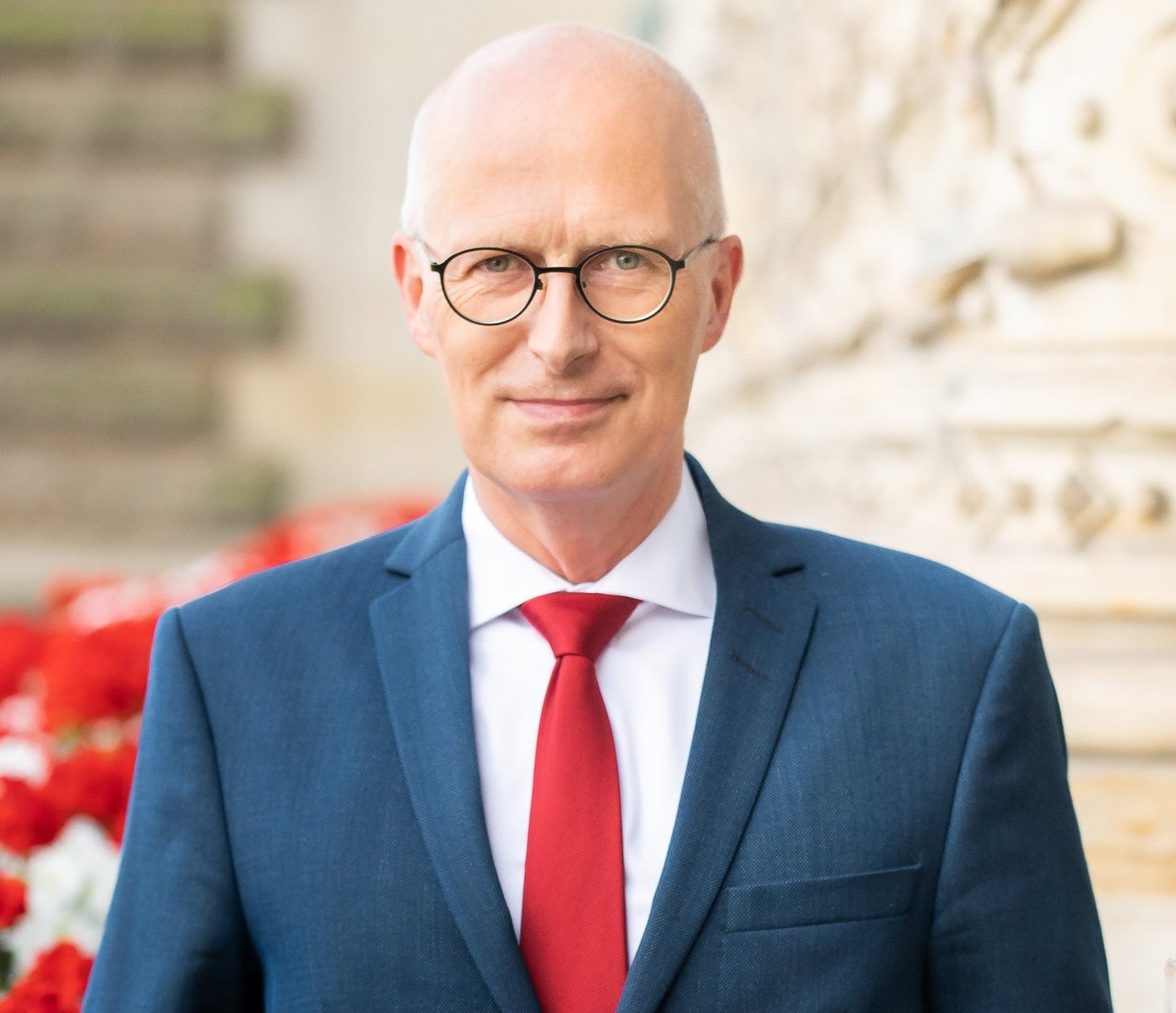 Dr. Peter Tschentscher Ein Mann in einem blauen Anzug mit roter Krawatte steht selbstbewusst vor einem historischen Gebäude, das mit kunstvollen Schnitzereien geschmückt ist. In der Nähe blühen leuchtend rote Blumen.