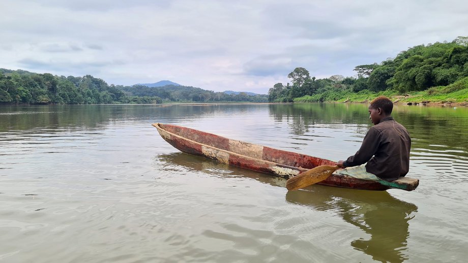 Gabun A young man sits in a narrow, brightly painted wooden boat on a calm river, surrounded by dense, green vegetation. His gaze is directed towards the water, with wooded hills stretching out in the background under a cloudy sky. The scene appears calm and close to nature.