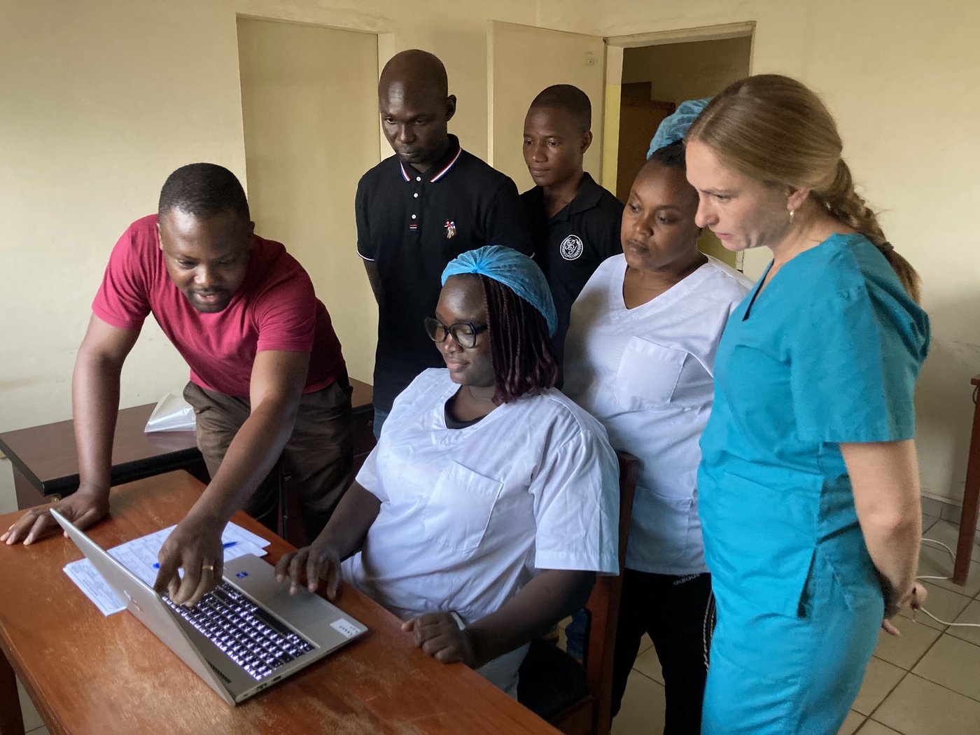 A lab group of five local staff from Benin plus one German trainer are looking at a laptop, one of them is pointing to the screen, highlighting something.