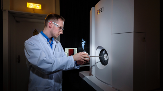 Krankheitserreger erforschen A person in a lab coat and safety glasses loads a sample into a large scientific instrument labeled FEI in a laboratory setting.