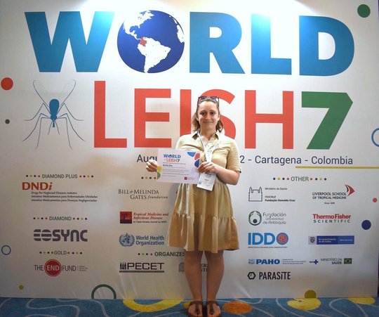 Poster Award for Annika Bea in Cartagena, Colombia. Eine Forscherin hält ein Urkunde in den Händen. Sie trägt ein knielanges, beigefarbenes Sommerkleid und eine in die Haare gesteckte Sonnenbrille. Hinter ihr ist ein großer Schriftzug "WorldLeish7" zu sehen, darunter mehrere Logos von verschiedenen Kooperationspartnern.