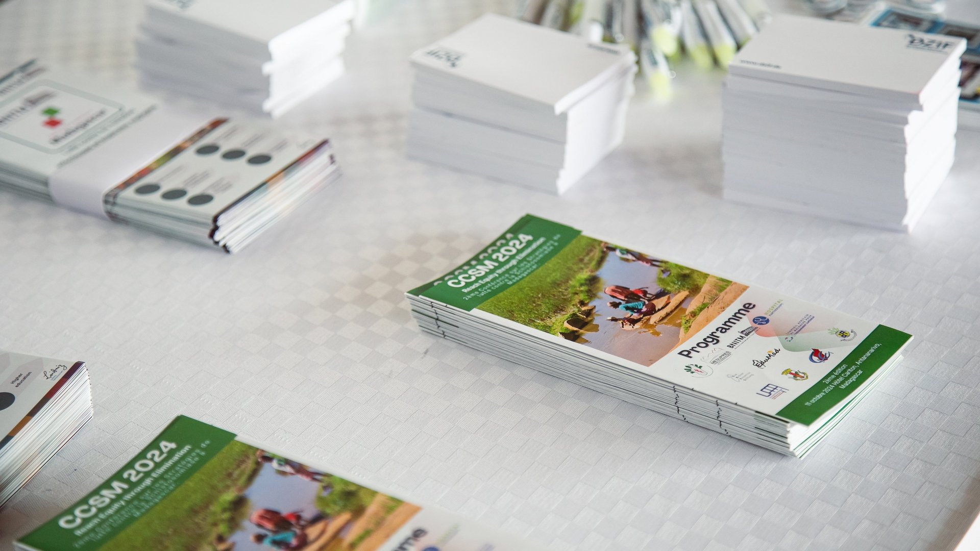 The image shows stacks of "CCSM 2024" brochures with a green banner and a photo of people near water, labeled "Programme," alongside other event materials like notepads and pens on a white tablecloth, suggesting a conference setup.