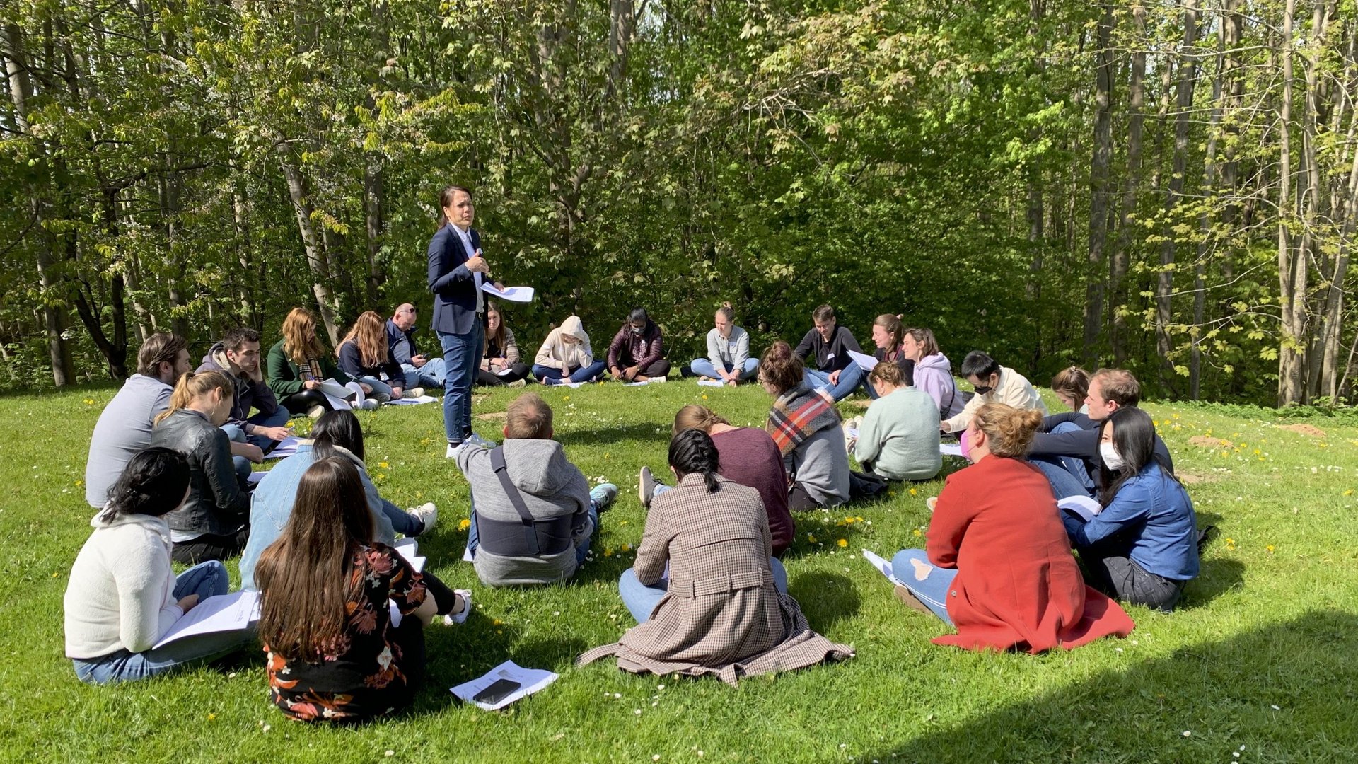 Eine Gruppe internationaler Teilnehmender eines Workshops sitzt in einem Kreis auf einer Wiese, in der Mitte steht eine Kursleiterin und erläutert Inhalte des Workshops.