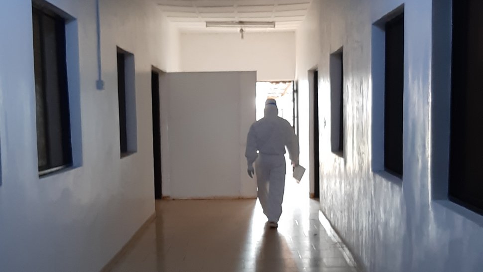 Lassafieber-Isolationsstation am ISTH Das Foto zeigt einen weißen, leeren Flur, mit Fenstern an den Seiten zu Behandlungsräumen. Am Ende des Gangs kommt eine Person in einem weißen Ganzkörperschutzanzug auf einen zu.
