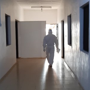 Lassafieber-Isolationsstation am ISTH Das Foto zeigt einen weißen, leeren Flur, mit Fenstern an den Seiten zu Behandlungsräumen. Am Ende des Gangs kommt eine Person in einem weißen Ganzkörperschutzanzug auf einen zu.