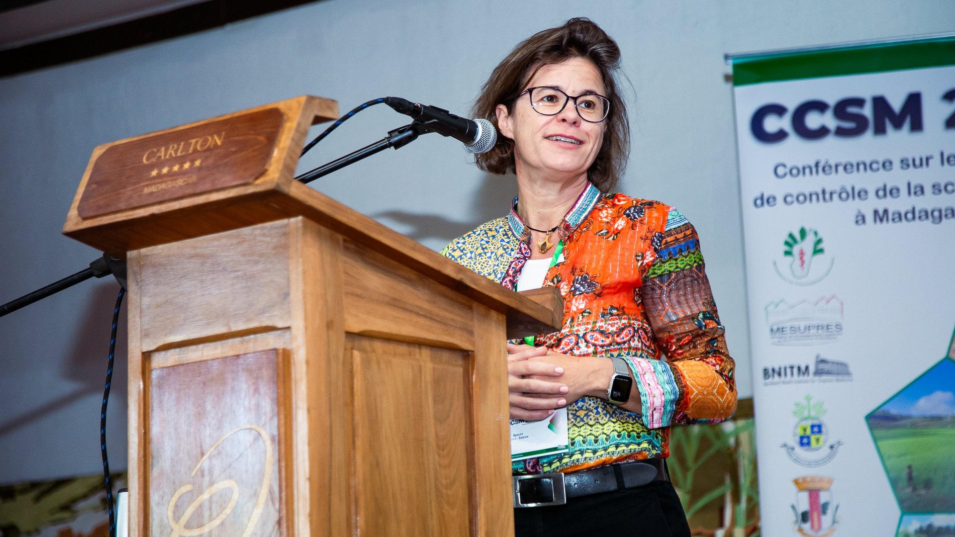 A woman speaking at the CCSM 2024 conference at a wooden podium, wearing a colorful blouse, with a CCSM 2024 conference banner in the background.