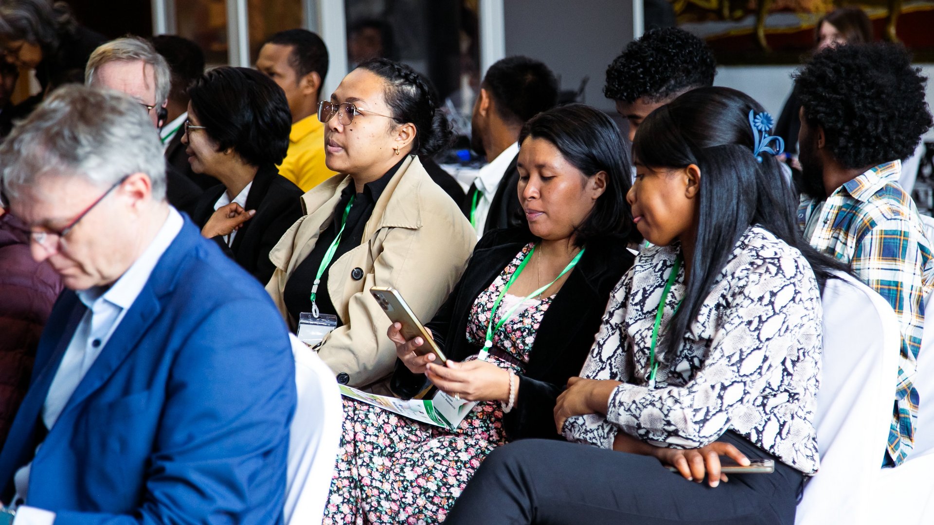 A group of CCSM 2024 attendees seated during a conference, some engaged with their phones and documents, wearing event badges with green lanyards.