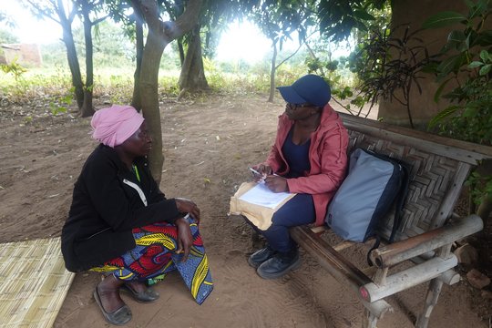 Interview being conducted with a traditional healer. Two women sit in front of each other. One woman is holding a pen and a notebook.