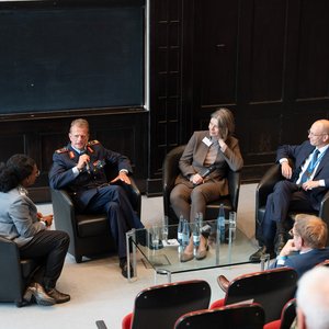 Podiumsdiskussion Das Foto zeigt vier Personen, die im Hörsaal auf vier dunklen Sesseln im Halbkreis sitzen.