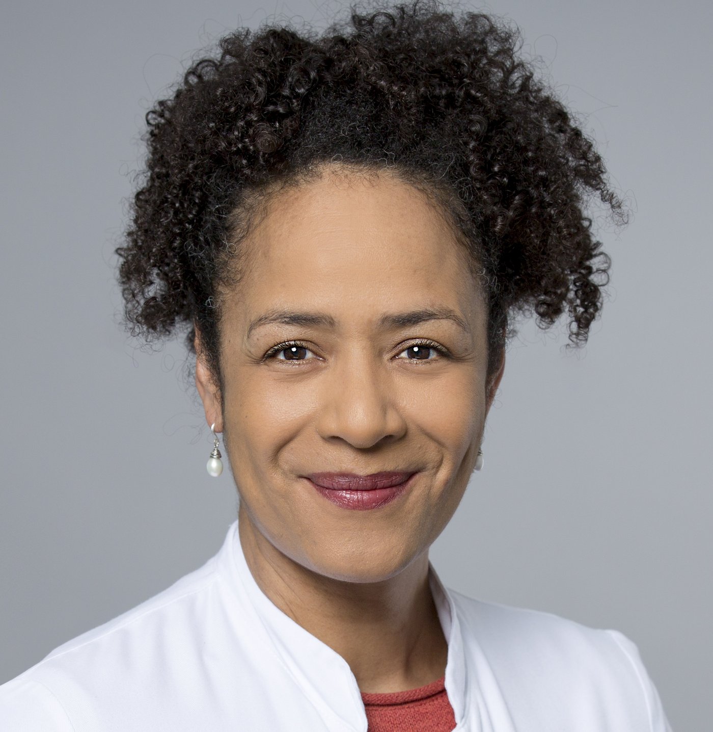 Prof. Dr. Marylyn Addo: Das Bild zeigt eine Ärztin in weißem Arztkittel mit lockigen schwarzen Haaren, die zu einem Zopf zurückgebunden sind und weißen Ohrringen.