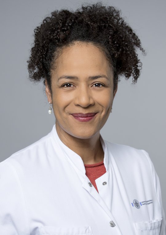 Prof. Dr Marylyn Addo Prof. Dr Marylyn Addo: The photo shows a physicist in a white coat with curly black hair tied back in a plait and white earrings.