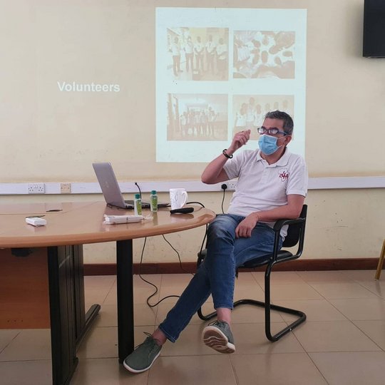 Training and capacity building during the hospital partnership workshop in Tanzania Zu sehen ist ein Mann, der vor einem an die Wand gebeamten Bild sitzend einen Vortrag hält. Man erkennt das Wort Volunteers auf der Projektion. Er trägt Mundschutz
