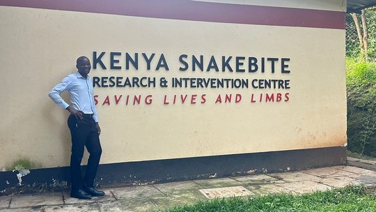Moses Aron in front of the sign of the K-SCRIC. The picture shows a man standing in front of a sign which says 'Kenya Snakebite Research & Intervention Centre - Saving Lives and Limbs'.