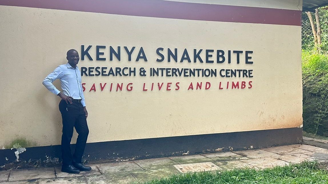 The picture shows a man standing in front of a sign which says 'Kenya Snakebite Research & Intervention Centre - Saving Lives and Limbs'.