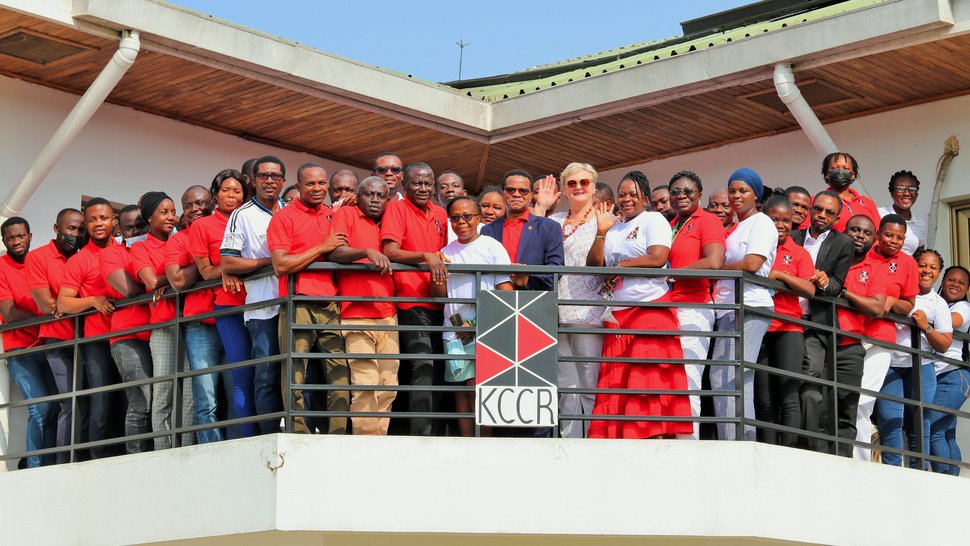 KCCR family photo KCCR employees in predominantly red T-shirts stand on the balcony of the institute, some waving. In the front, in the middle, stand the scientific director Prof. Phillips and the blonde managing director Ingrid Sobel.