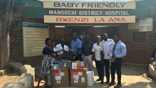 Handover of ultrasound machines to Mangochi District Hospital The picture shows nine people standing in front of a hospital. In front of them are packages and an ultrasound machine is visible which is being held by two people.