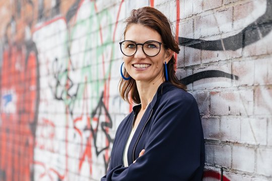 Prof. Dr. Cornelia Betsch Foto von Cornelia Betsch vor einer Graffiti-Backsteinwand. Sie trägt ein dunkelblaues Kostüm, schulterlange dunkelbraune Haare, eine markante dunkel umrahmte Brille und blickt freundlich in die Kamera.